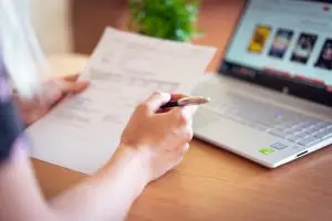 Close-up of a hand holding a pen and paper near a laptop