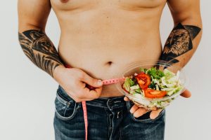 Tattooed man measuring waist while holding fresh salad